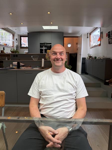 A bald man with a playful expression sits at a glass table, looking directly at the camera